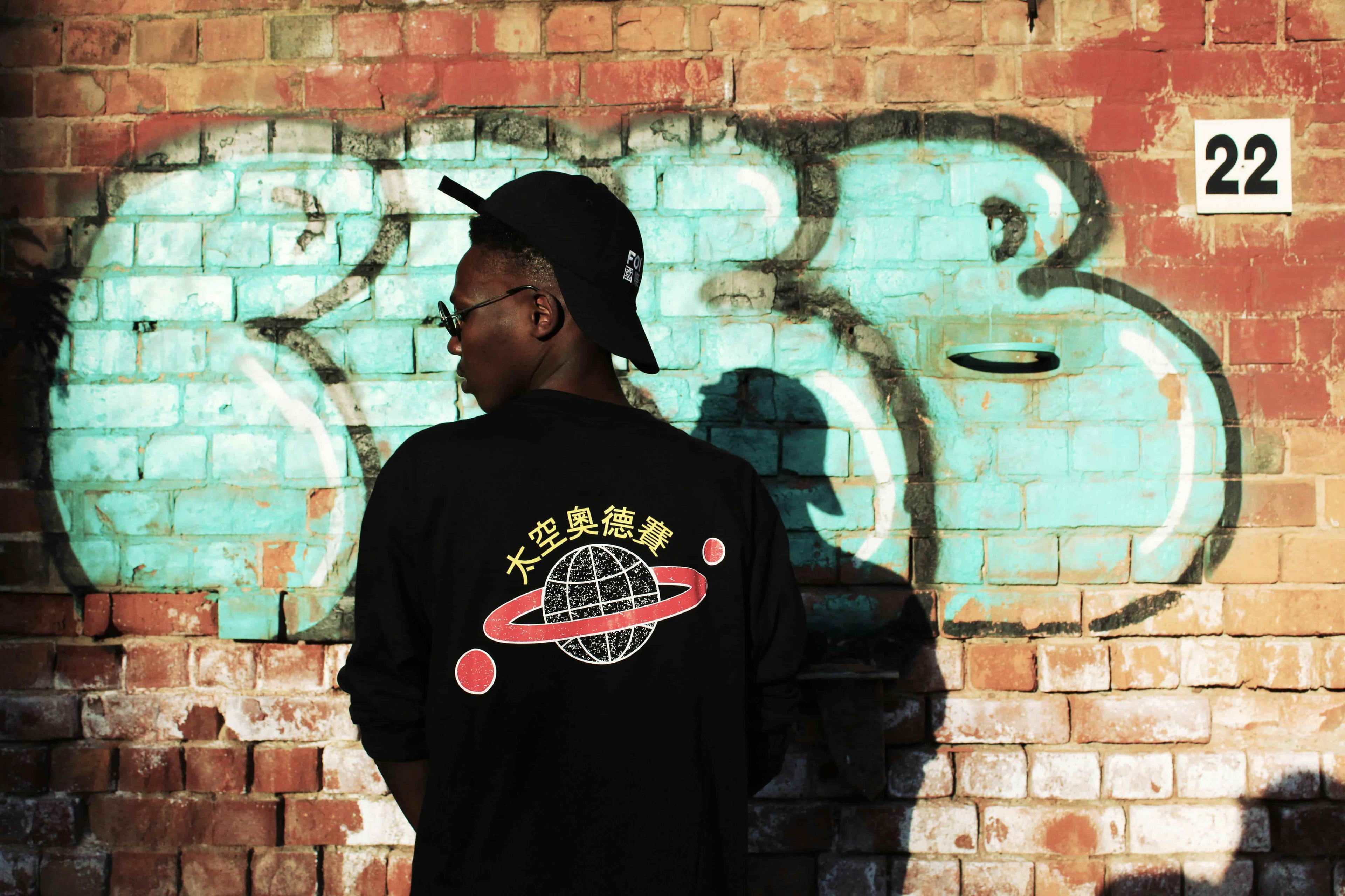 Person wearing a black hoodie with a graphic design in front of a graffiti-covered wall.